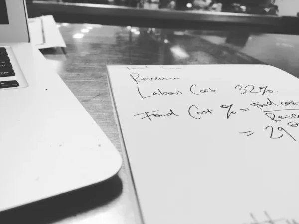 Handwritten notes working out labour cost and food cost percentages beside a laptop